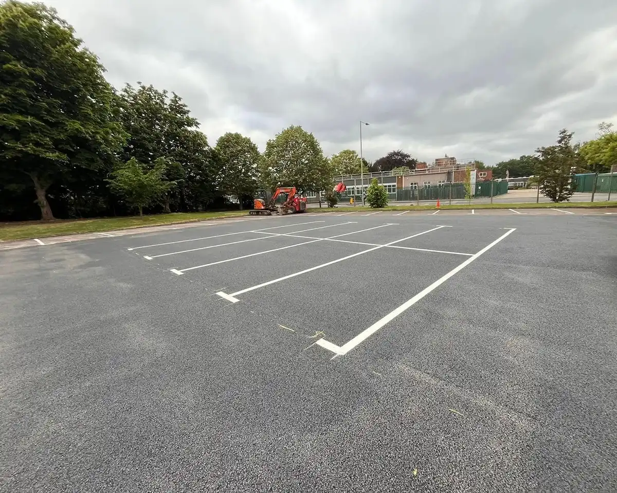 Empty car park with white-lined spaces, a red vehicle parked nearby, and trees in the background under a cloudy sky.