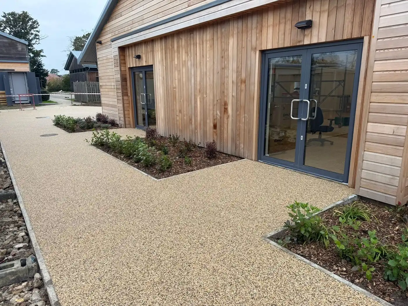 Entrance path to a modern building, featuring a textured gravel surface and landscaped areas with small shrubs and plants.