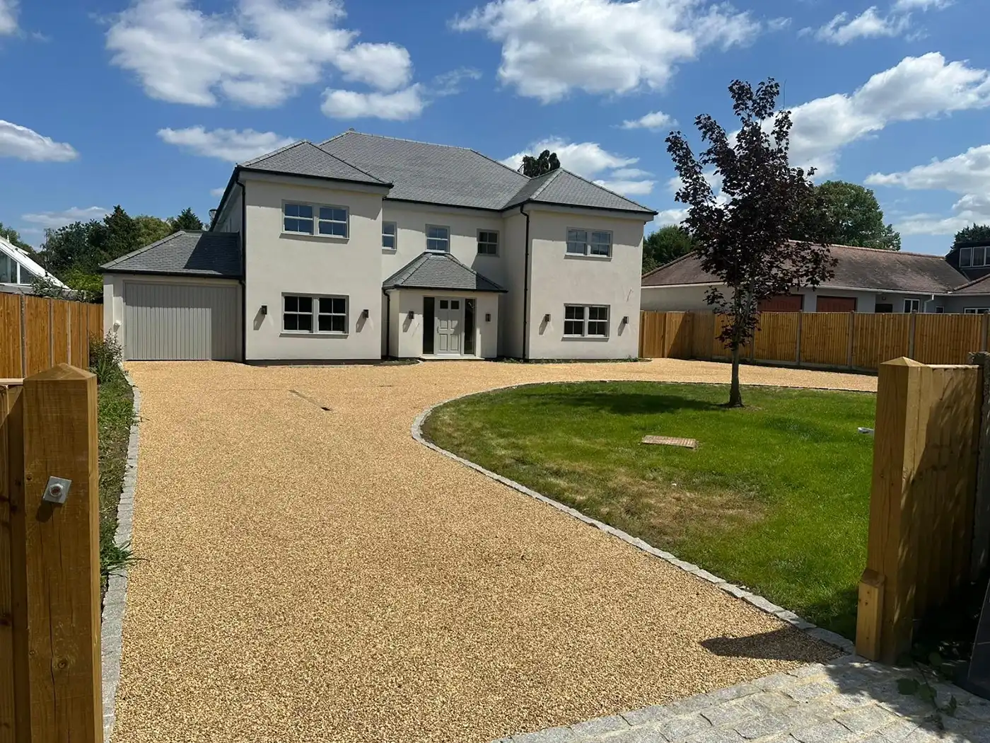 Modern two-story house with a grey roof, surrounded by a circular gravel driveway and a green lawn, under a blue sky with clouds.